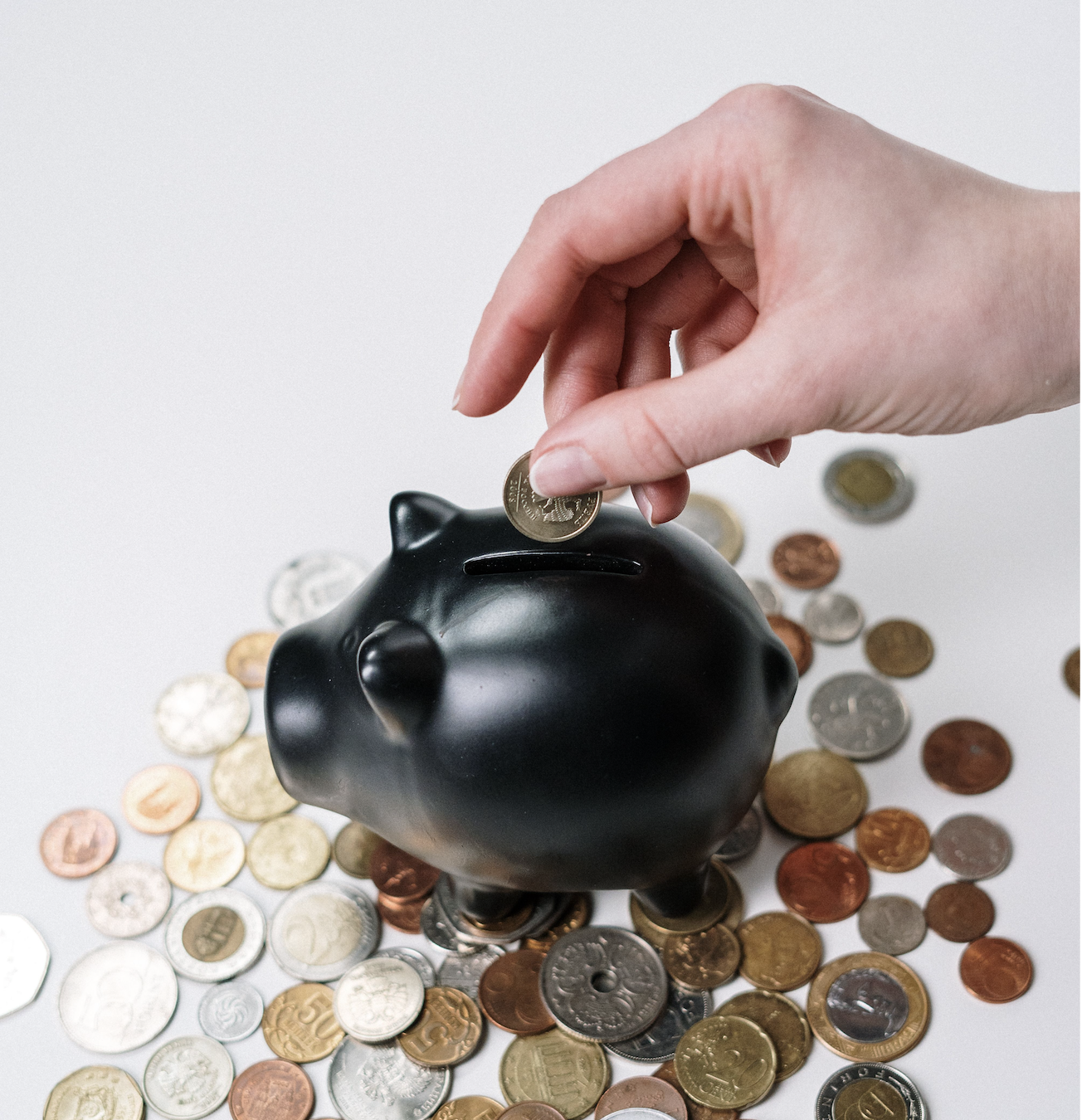 Black ceramic piggy bank sitting on a pile of coins of different currencies against a white background with a white person's hand putting a coin into the piggy bank