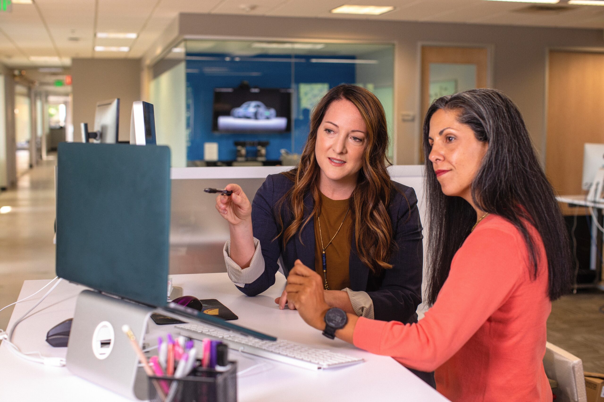 two women sitting in front of a computer monitor working together in an open office