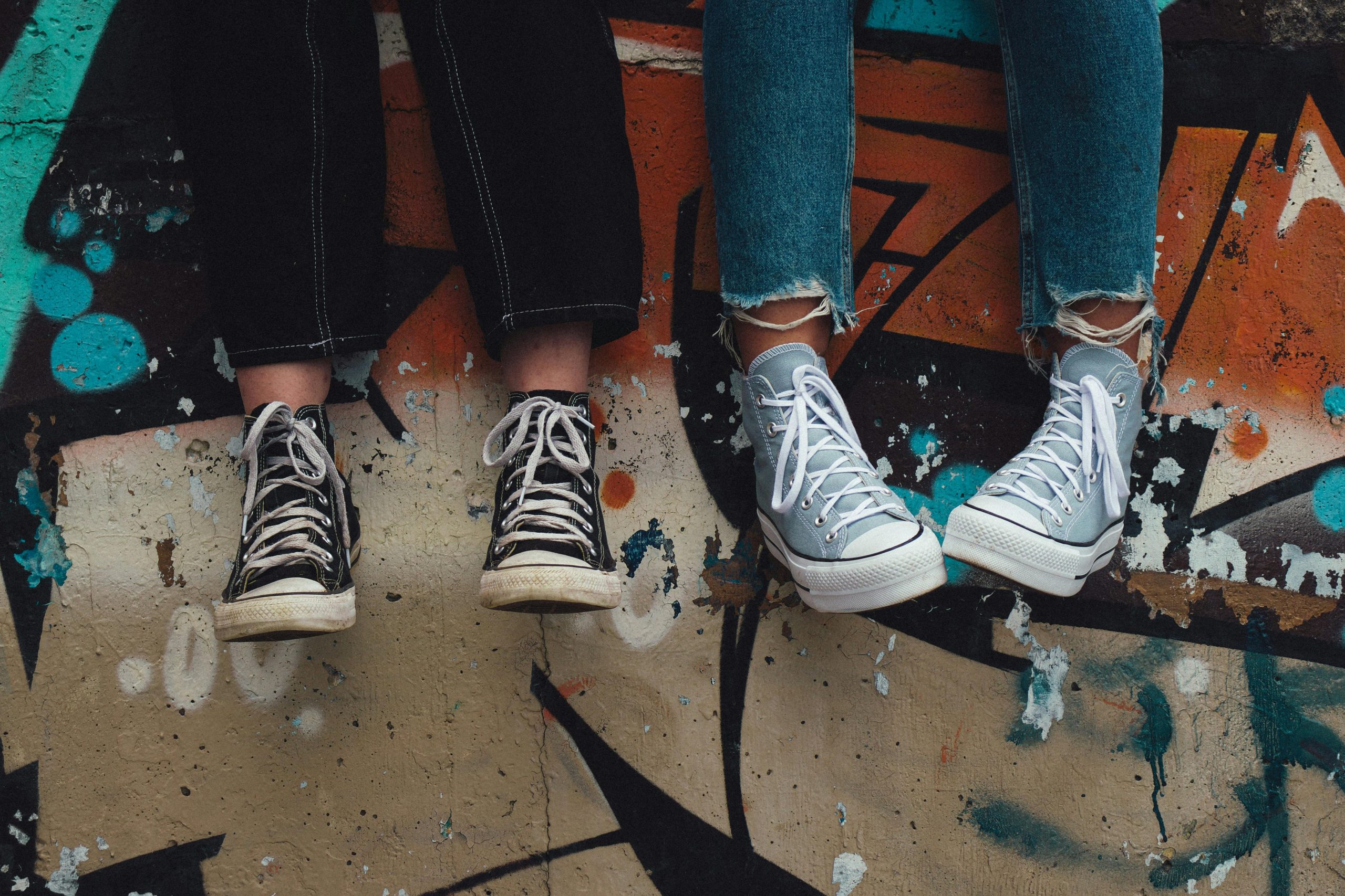 Two pairs of legs and feet - one wearing black jeans and black high-top Chuck Taylors, the other wearing blue jeans and grayish blue high-top Chuck Taylors - against a background of a wall with graffiti in shades of orange and blue