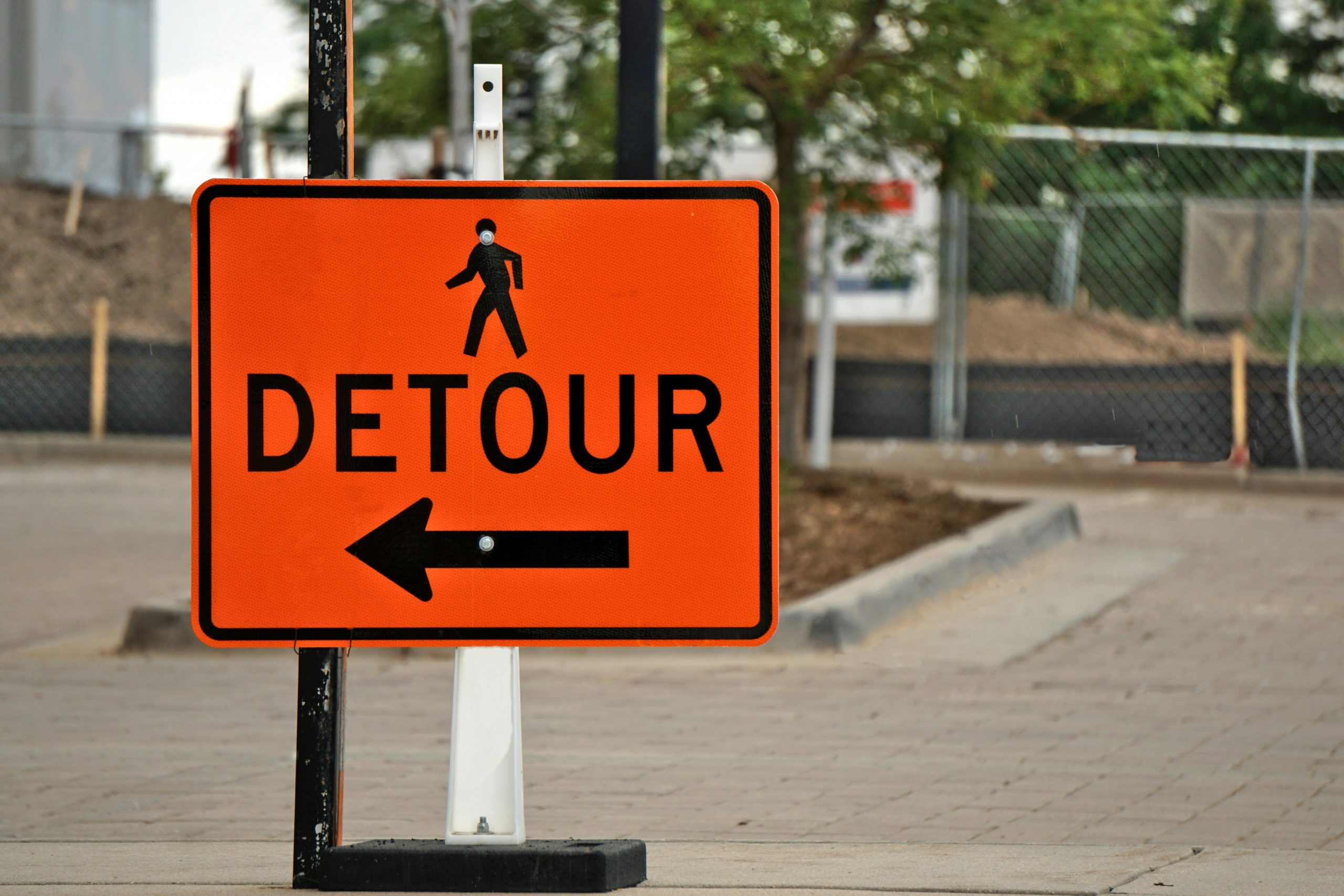 Black on orange road work style sign that reads DETOUR with a walking stick figure and an arrow that points left against an out of focus background of a sidewalk with a tree box and chain link fence